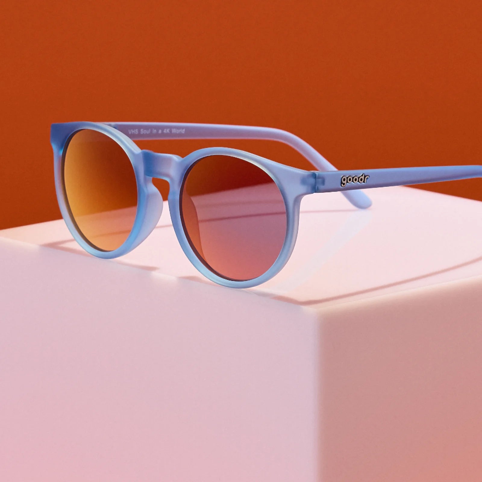 Product showcase of dusty blue circle-shaped sunglasses with red reflective lenses. 